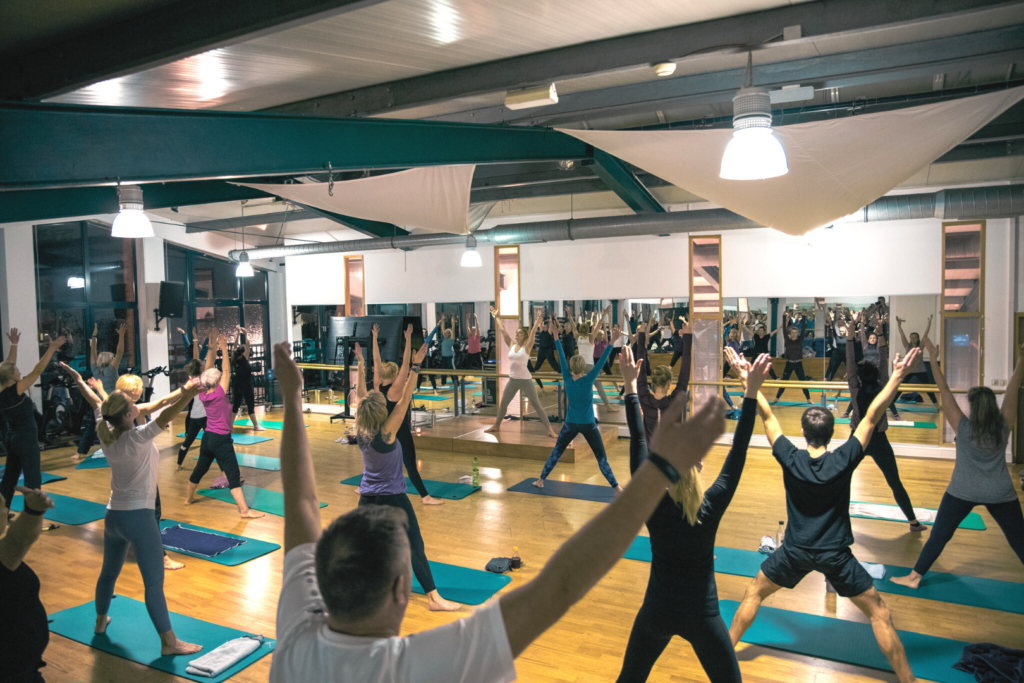 13 Menschen die gemeinsam in einem Raum an einem Yoga Kurs teilnehmen
